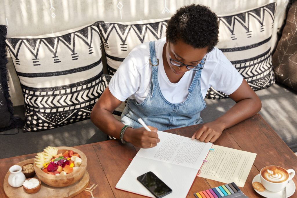 top view of attractive stylish black woman writes review of book in notepad